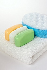 Three soap bars of same size but different colors (orange, white and green) and textures, on a white fluffy towel folded in half and next to a double sided blue and white porous sponge. The bars have a slightly concave shape with curved corners.