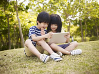 little asian girl and boy sitting on grass using digital tablet outdoors in a park.